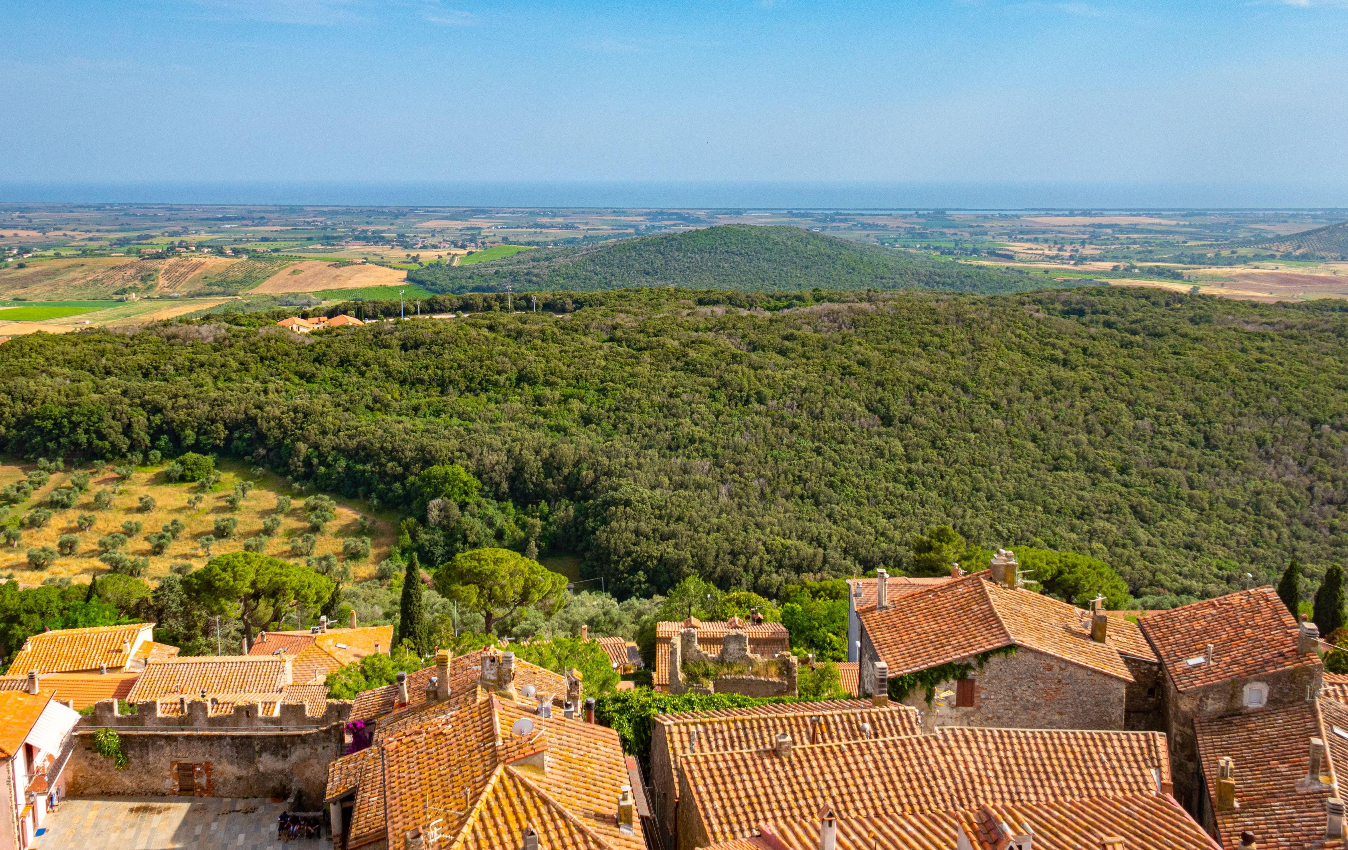 Tuscan landscape viewed from a hilltop, showing terracotta rooftops, woods, and the sea in the distance.