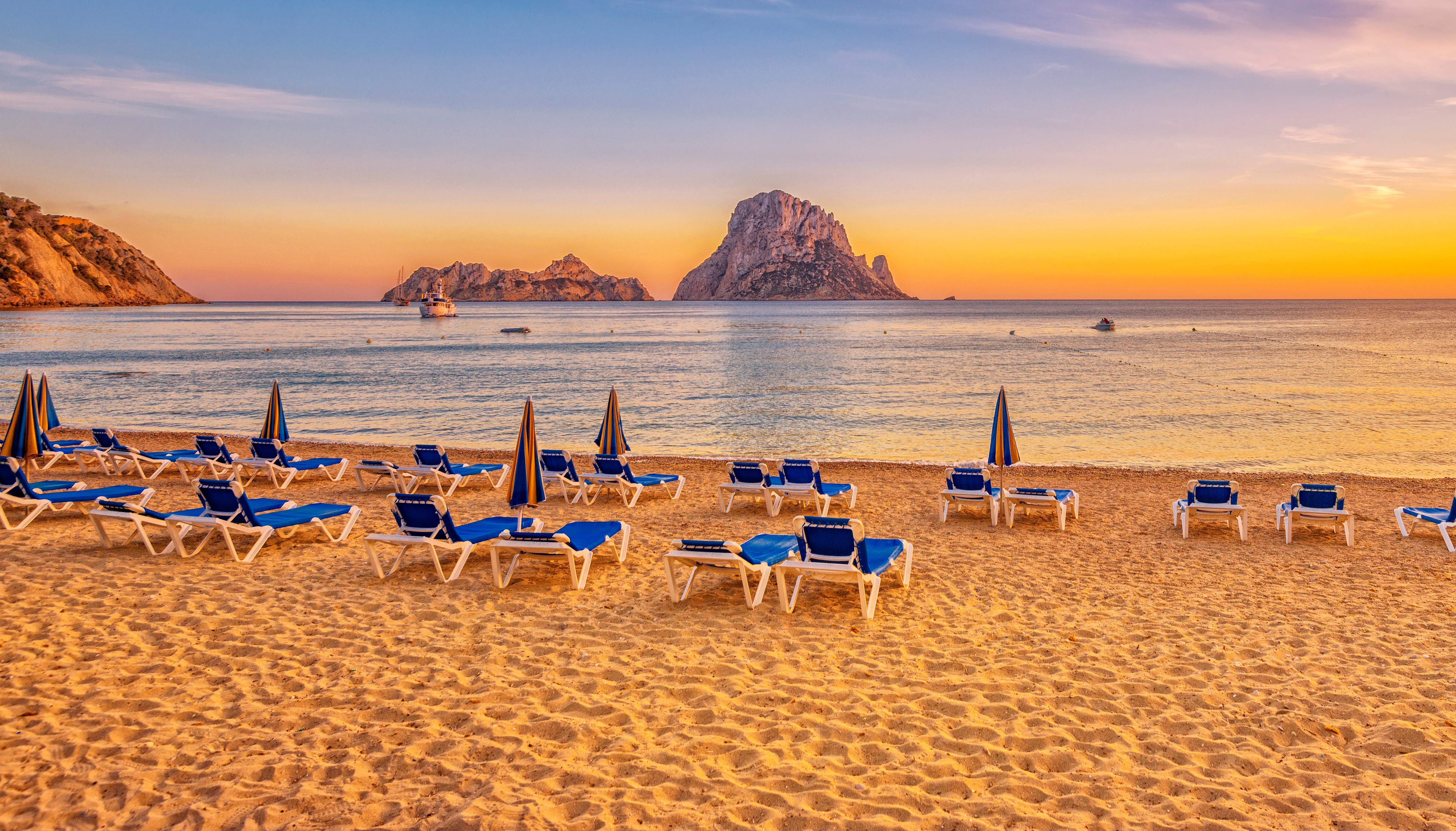 Sunset over Cala d'Hort beach in Ibiza, Spain, with lounge chairs and boats.